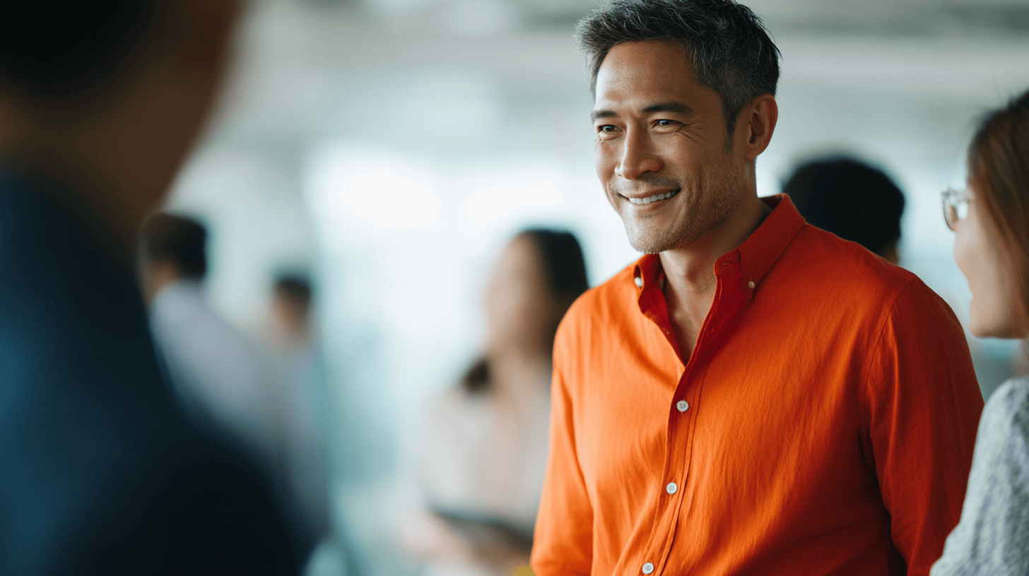 Smiling man in an orange shirt engages in conversation with blurred figures in a bright, modern setting.