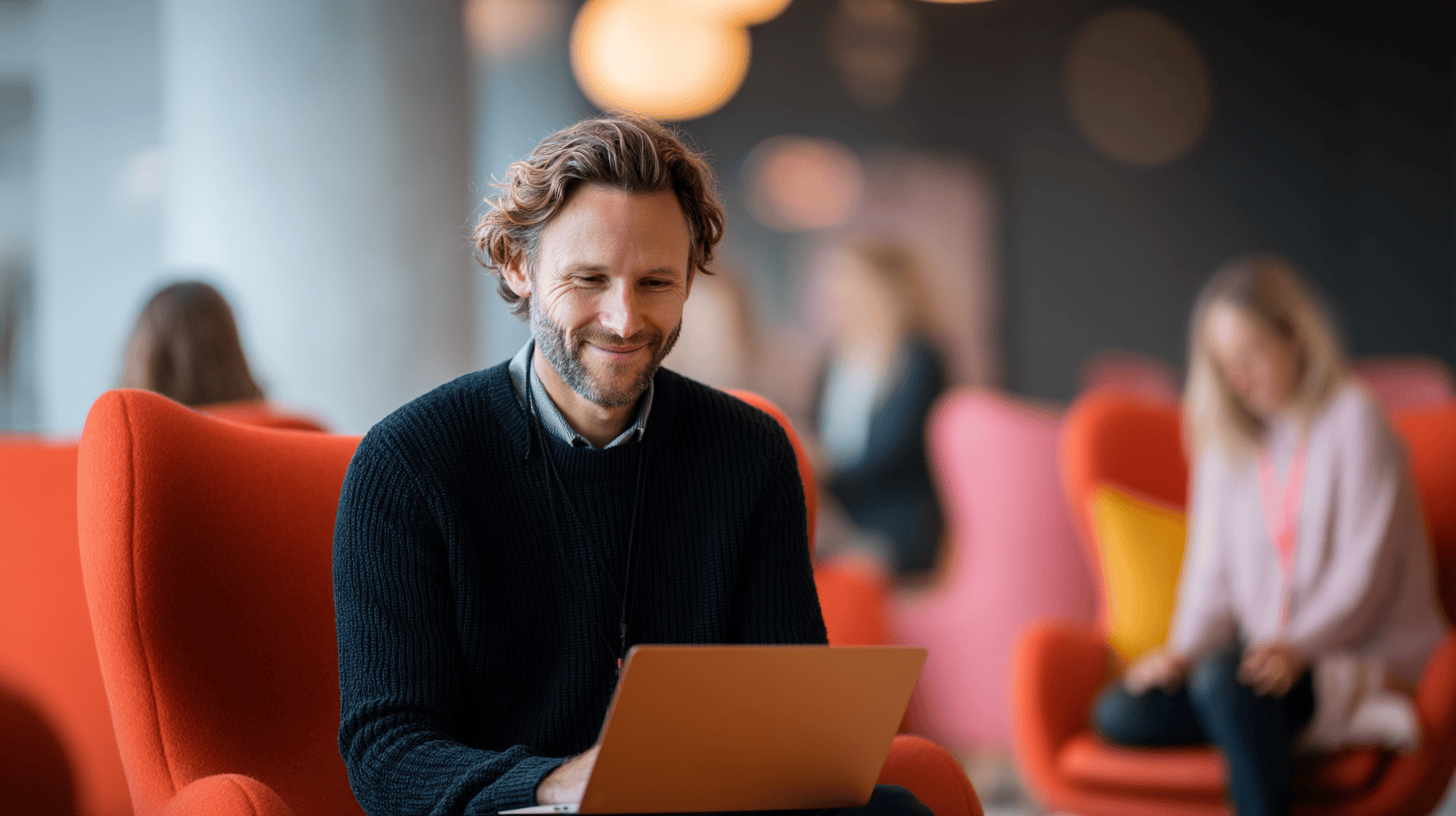 A man in a black sweater smiles at a laptop in a modern, colorful lounge with other people in the background.