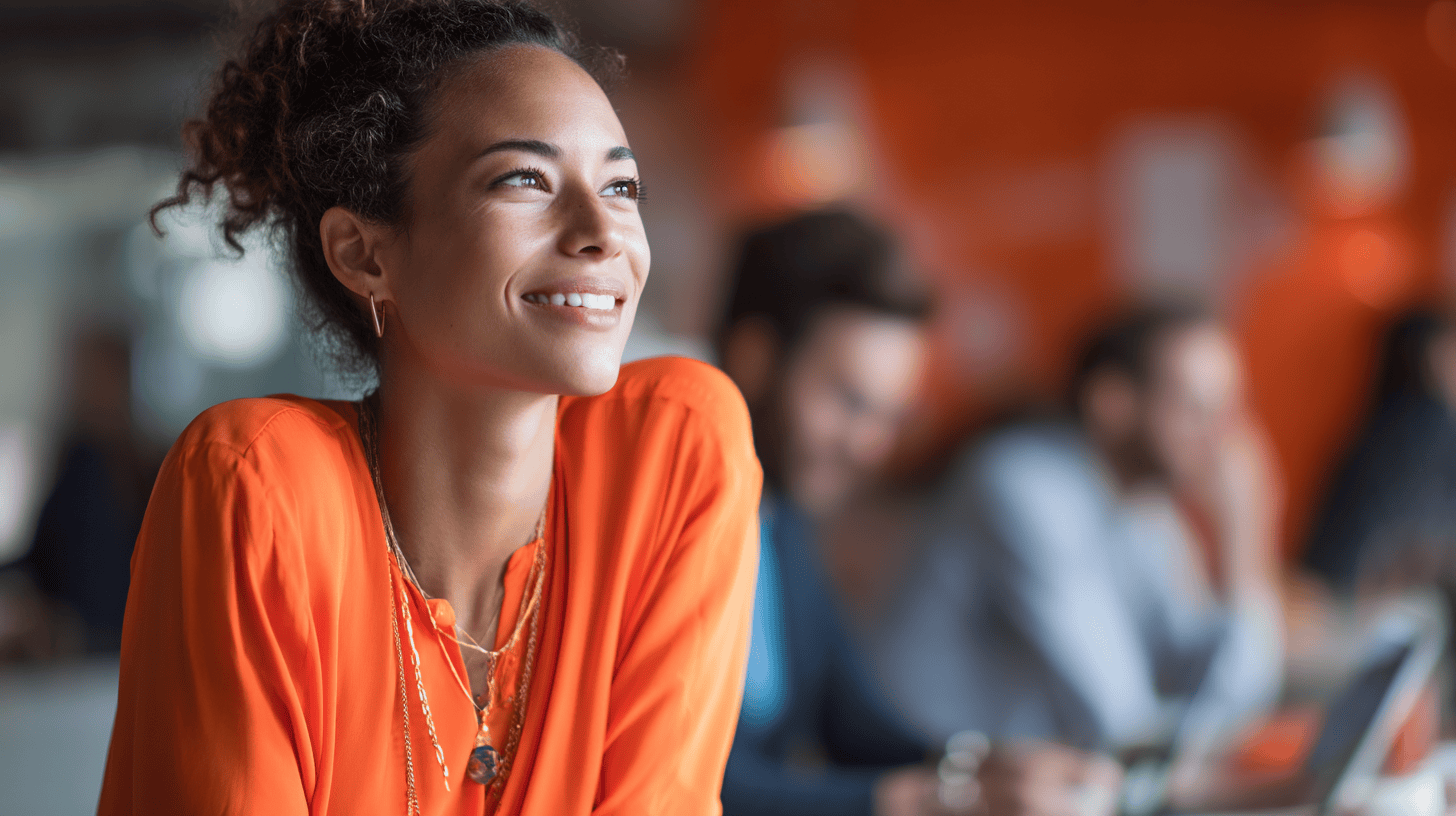 A smiling woman in an orange top is seated at a café, with two blurred individuals working in the background.