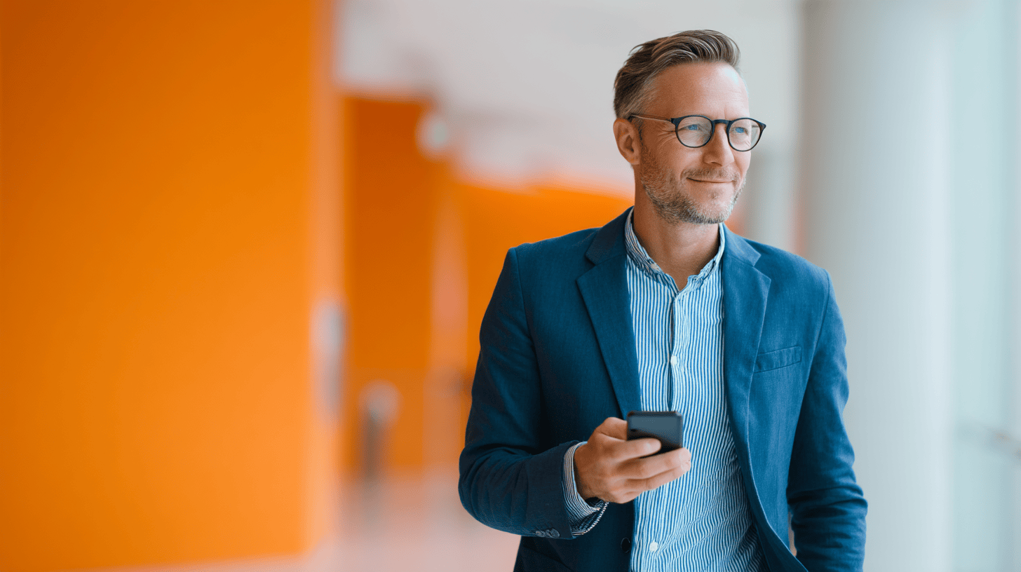 A smiling man in a blue blazer and glasses holds a smartphone in a brightly lit corridor with orange walls.