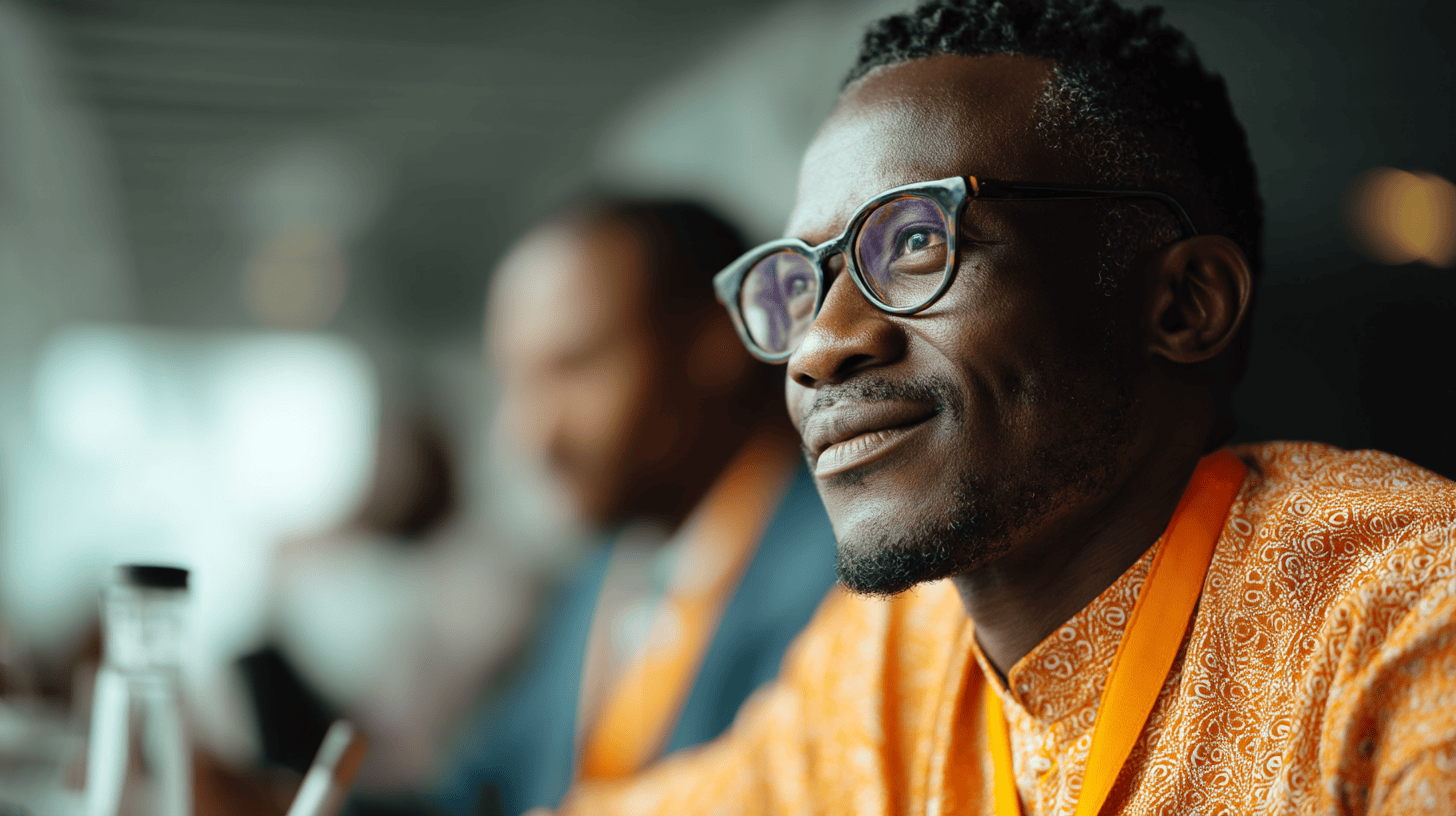 A man in glasses smiles while wearing an orange patterned shirt, with a blurred background of an event or conference.
