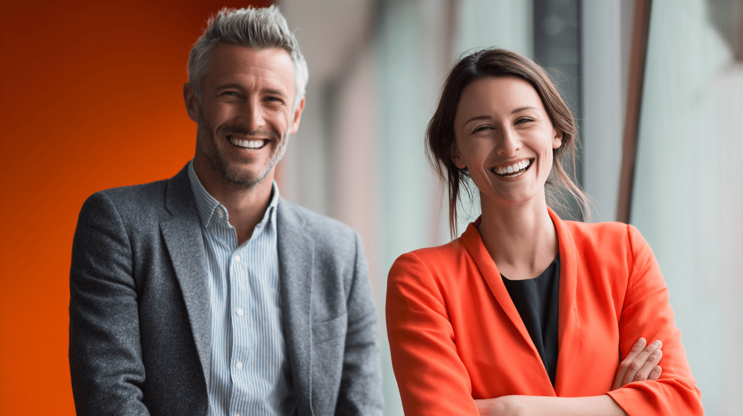 A smiling man in a gray blazer and a woman in an orange blazer pose together against an orange background.