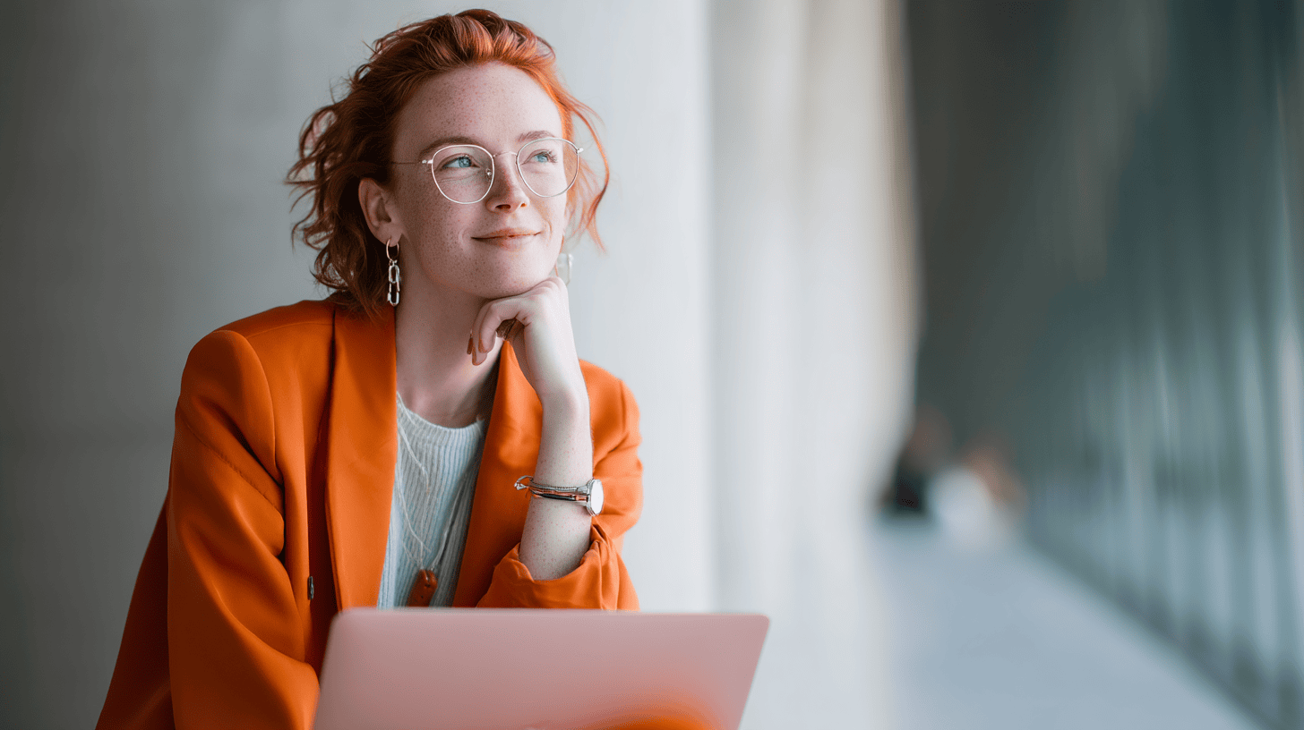 A woman with curly red hair and glasses wears an orange blazer, smiling while resting her chin on her hand, with a laptop in front.