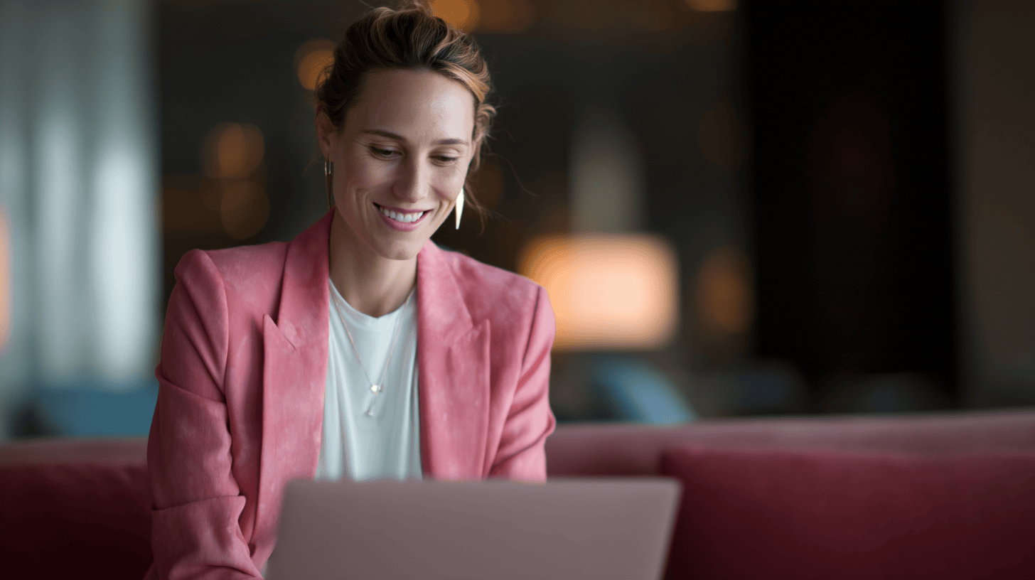 A smiling woman in a pink blazer sits on a couch, engaging with a laptop in a softly lit modern setting.