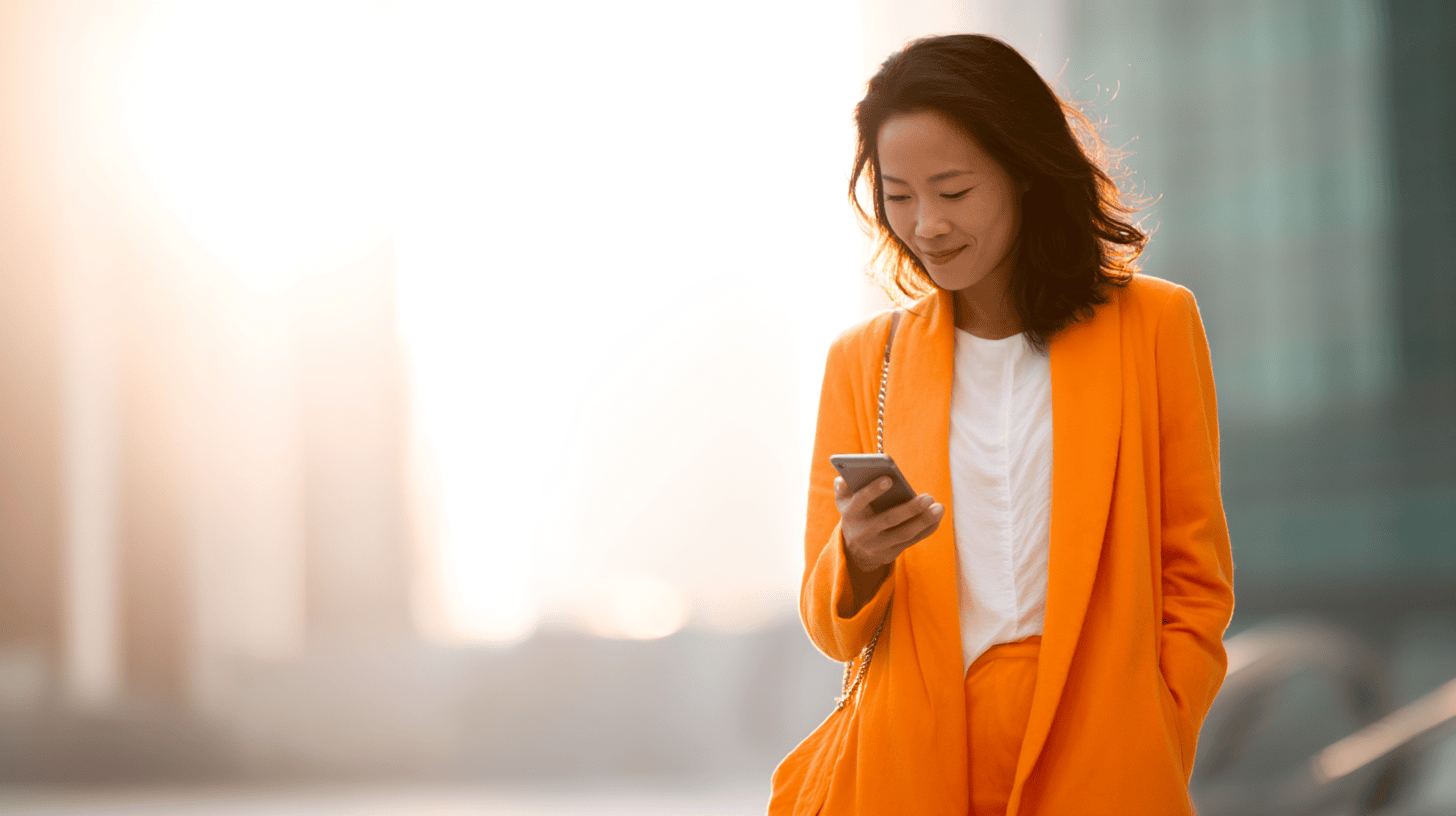 A woman in a bright orange blazer checks her phone, walking in a sunlit urban setting. Soft background blur enhances focus.