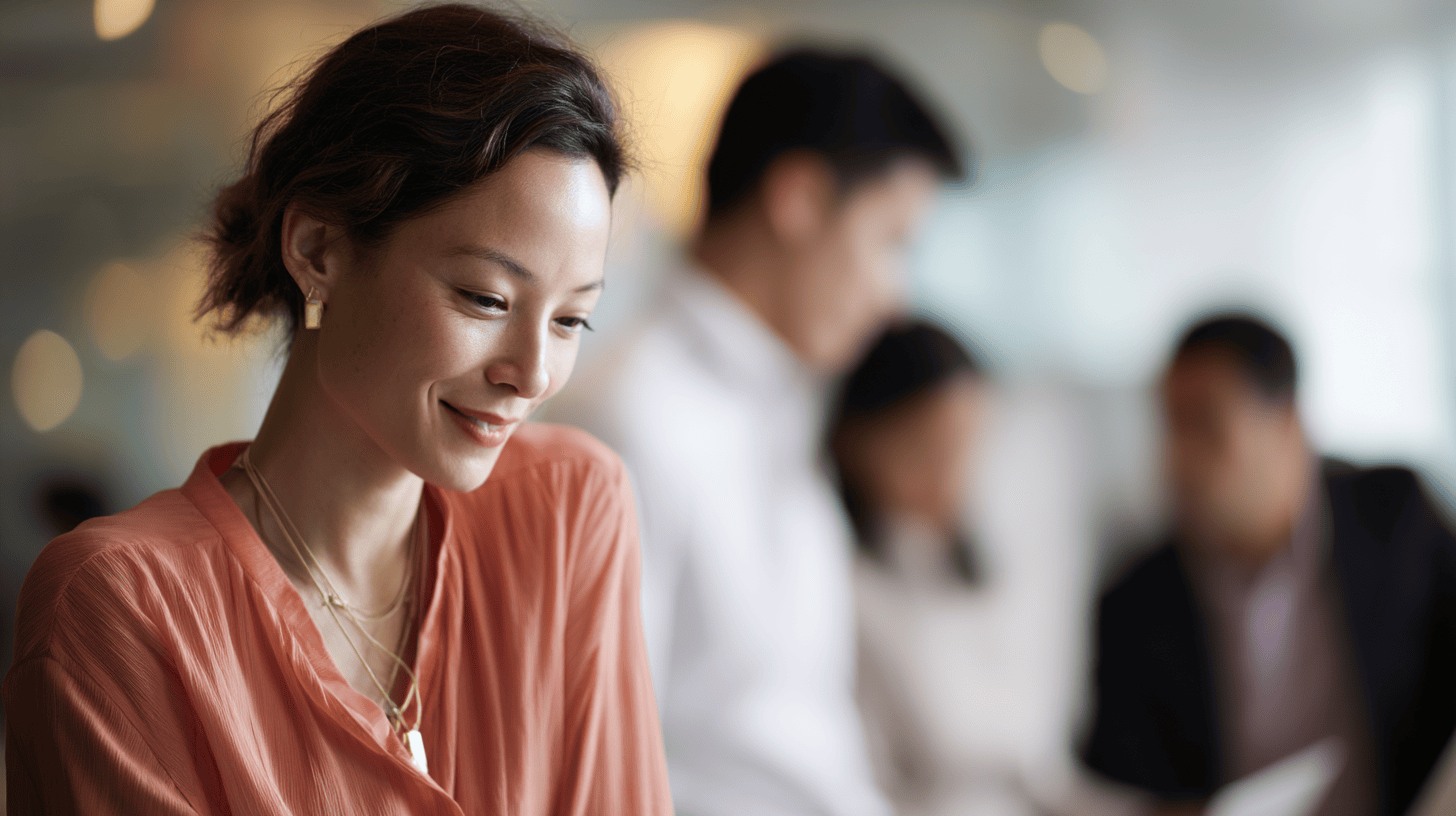 A woman in an orange blouse smiles while looking down, with blurred figures of people in a professional setting behind her.