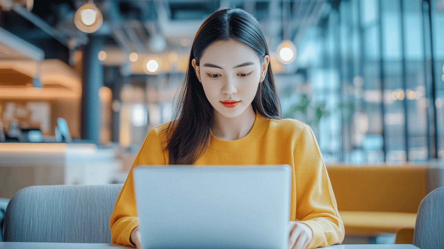 A young woman in a yellow sweater sits at a table, focused on her laptop in a modern, brightly lit space.