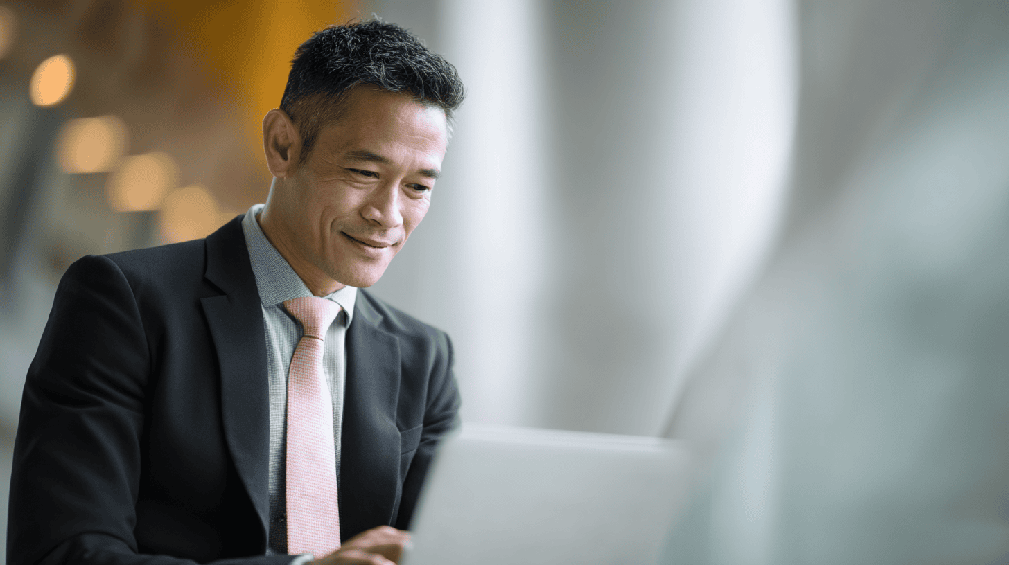 A man in a suit and pink tie smiles while working on a laptop in a bright, modern setting.