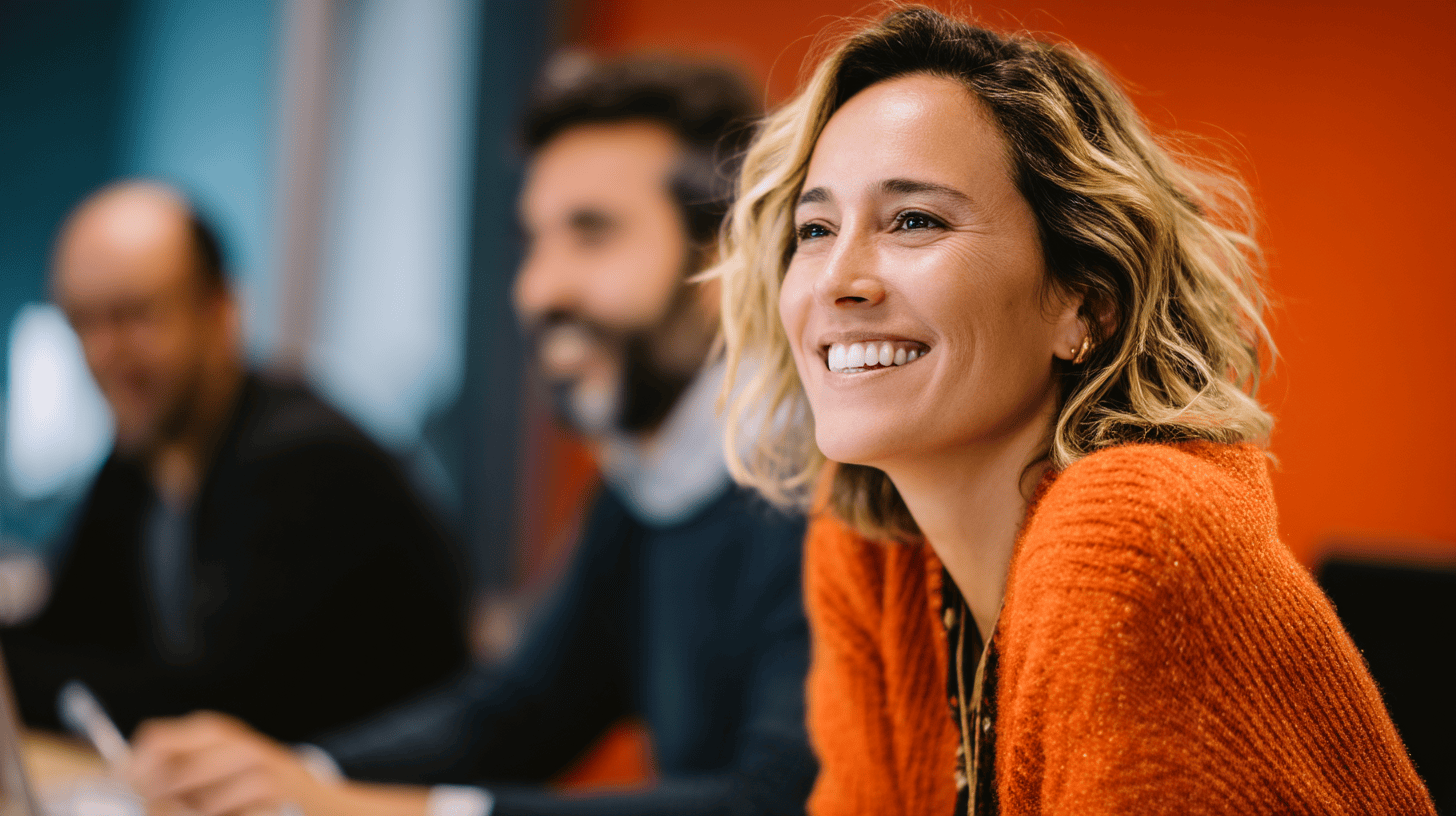 Smiling woman in an orange sweater at a table, with two people blurred in the background, against an orange wall.