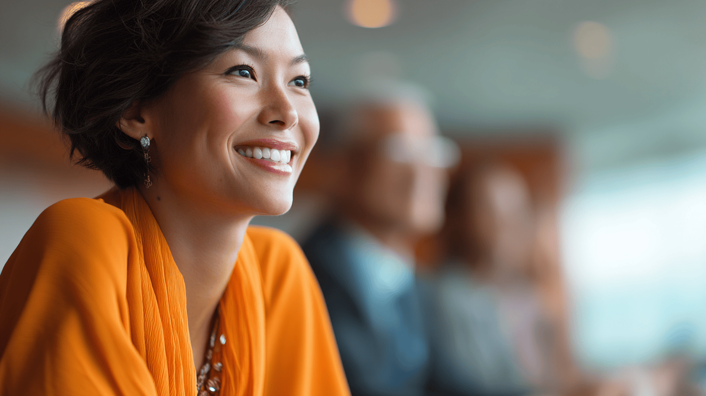 A woman in an orange blouse smiles brightly at a business meeting, with blurred colleagues in the background.