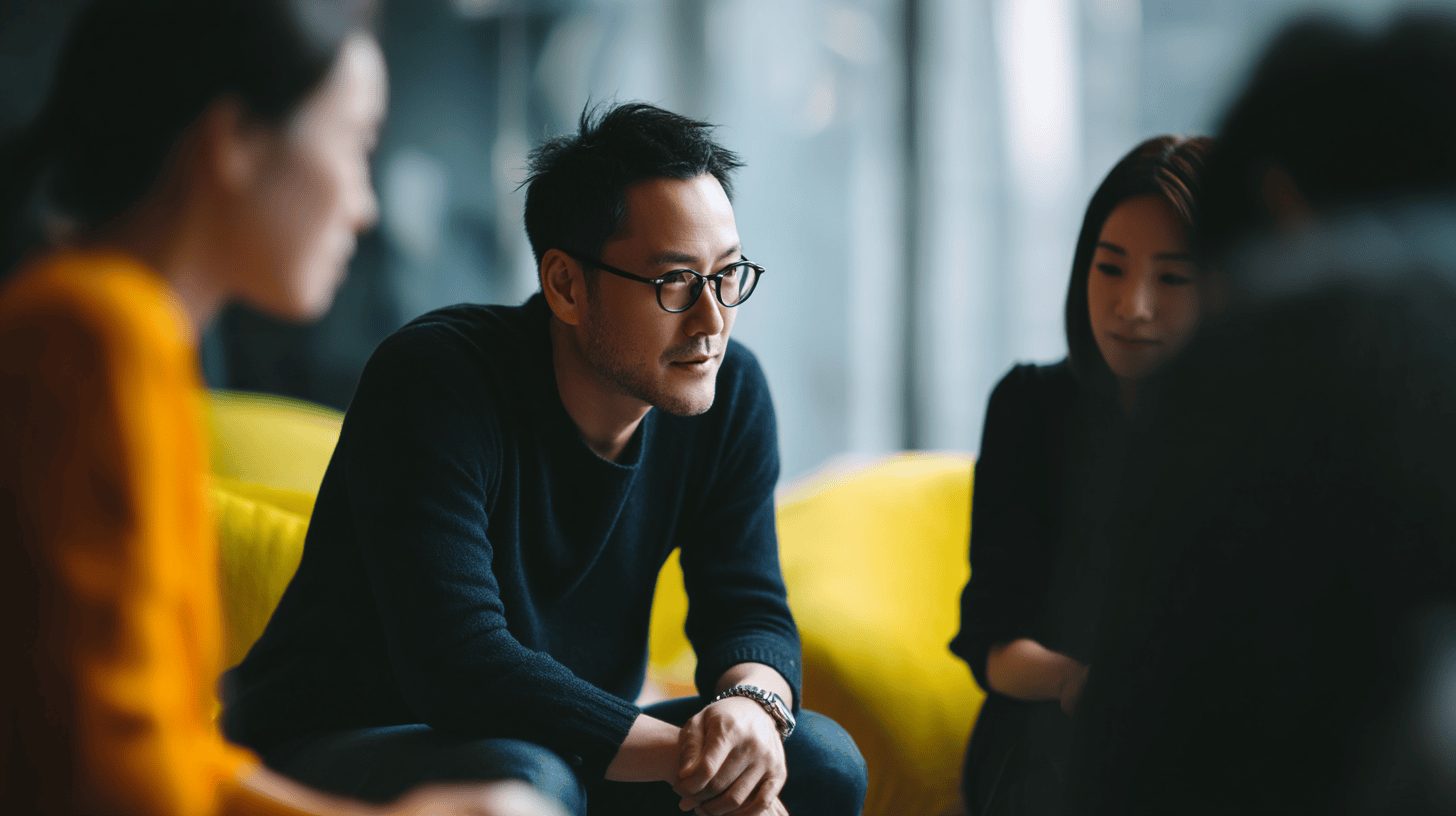 A group discussion with a man in a black sweater and glasses, sitting on a yellow couch with two blurred individuals.