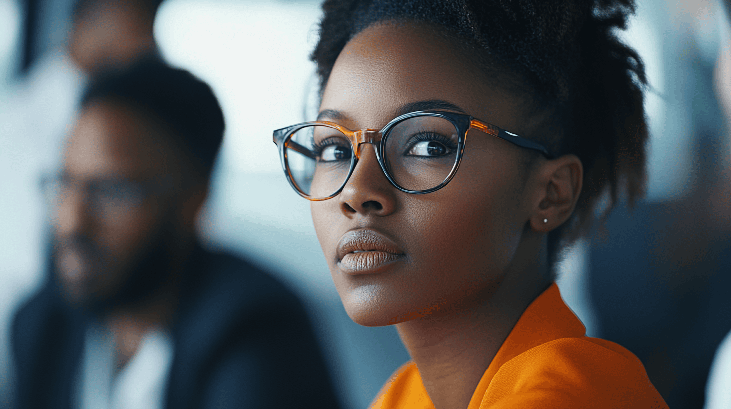 A woman with glasses and curly hair gazes thoughtfully, wearing an orange blouse; blurred figures are seen in the background.