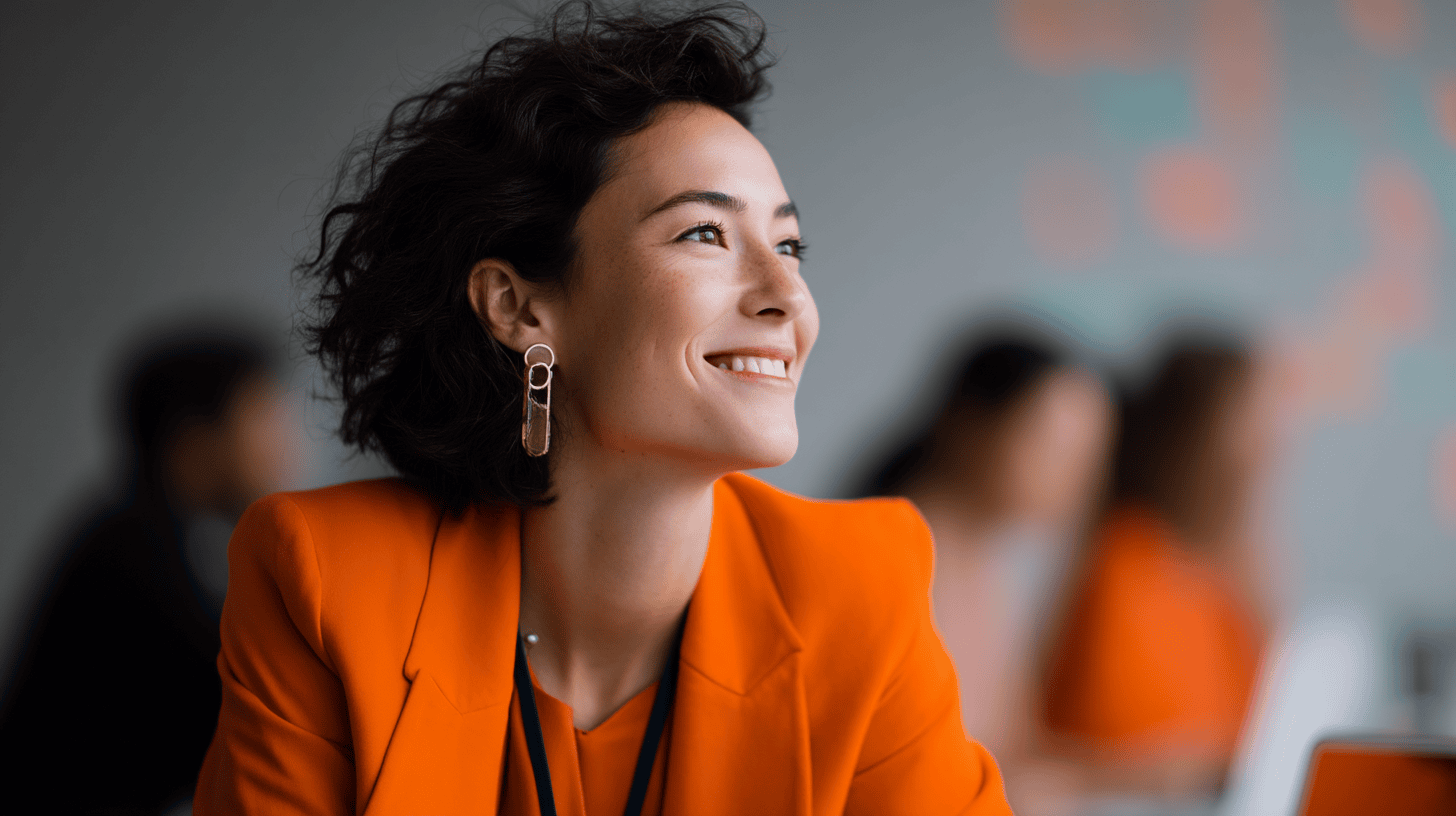 A woman with curly hair smiles, wearing an orange blazer and statement earrings, in a soft-focus office setting.