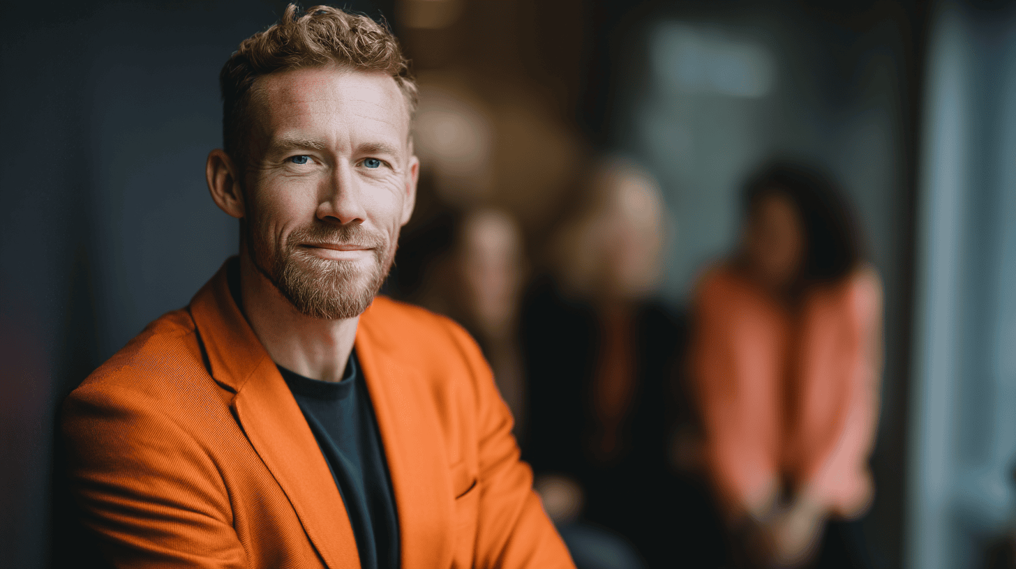 A man in an orange blazer smiles confidently at the camera, with blurred figures in the background.