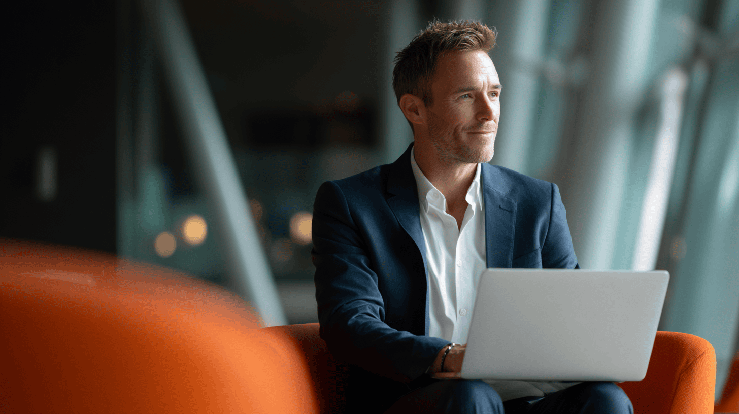 A man in a dark suit sits on an orange chair, looking thoughtfully at a laptop in a modern, bright space.