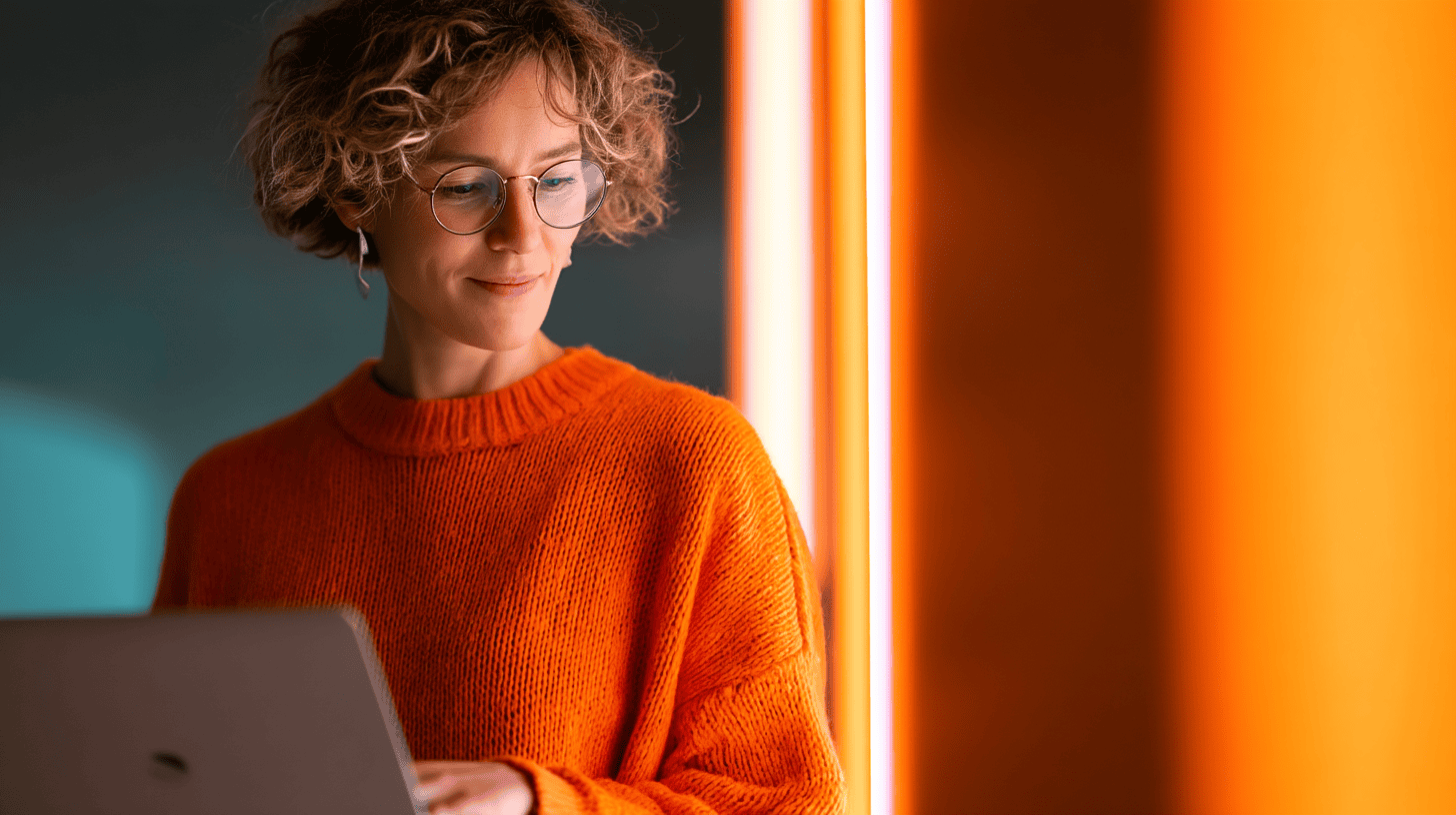 A woman with curly hair wears glasses and an orange sweater, working on a laptop near colorful lighting.