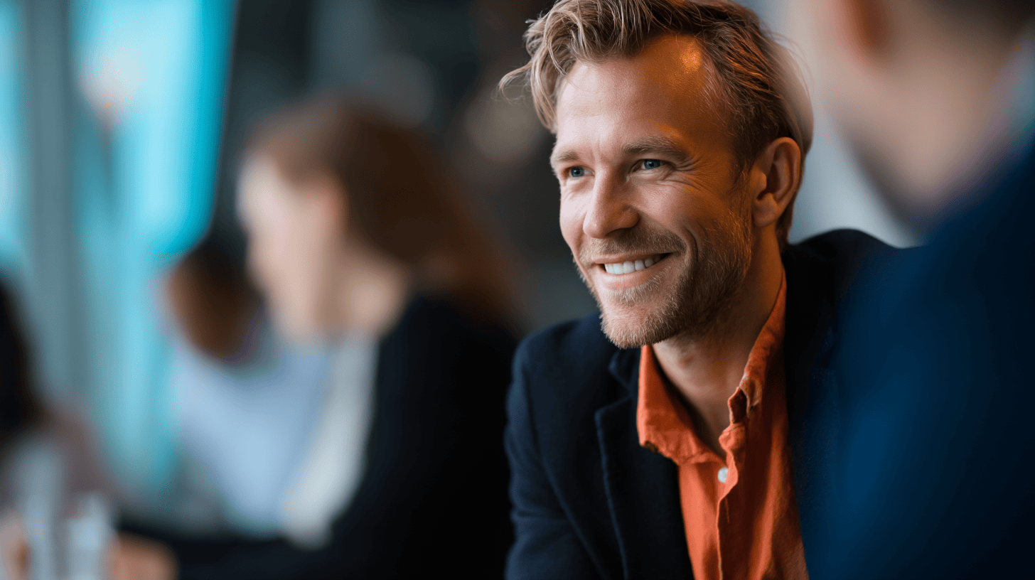 A smiling man with light hair and a beard wears a black blazer and orange shirt, sitting with blurred people in background.