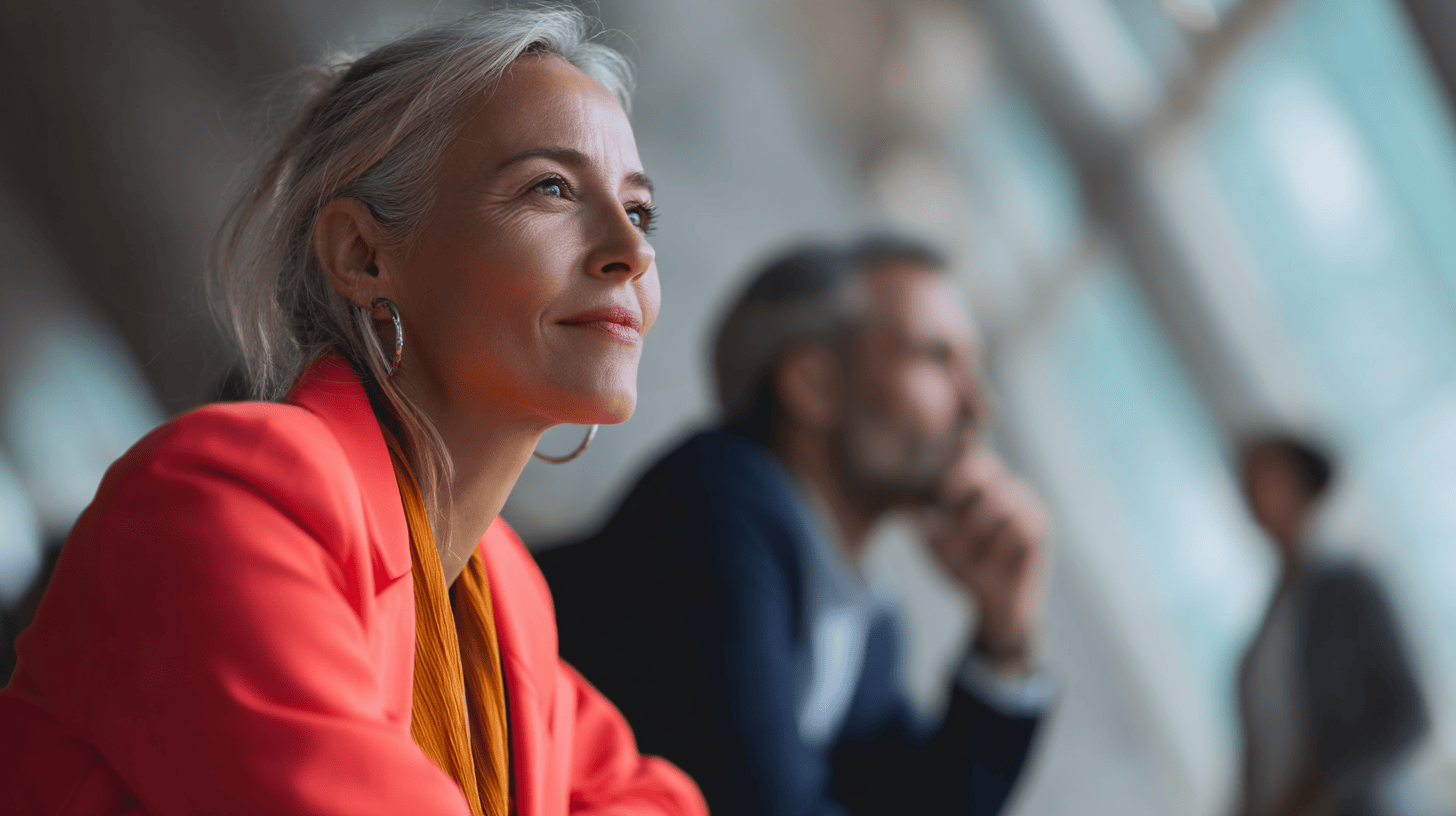 A woman in a bright orange blazer gazes thoughtfully, with a blurred man and another figure in the background.