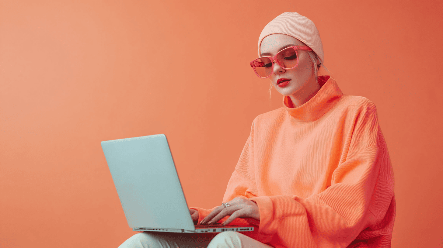 A person in a bright orange sweater and pink sunglasses sits with a laptop against an orange background.