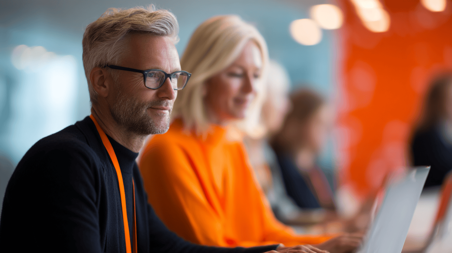 A man with glasses and a woman in an orange sweater focus on laptops in a modern, brightly lit office setting.