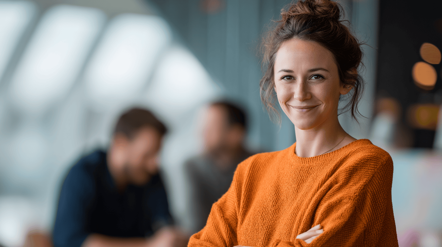A woman in an orange sweater smiles, with blurred figures of people in the background, inside a modern space.
