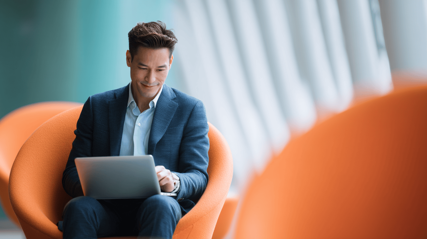 A man in a suit sits on an orange chair, focused on a laptop in a bright, modern indoor setting.
