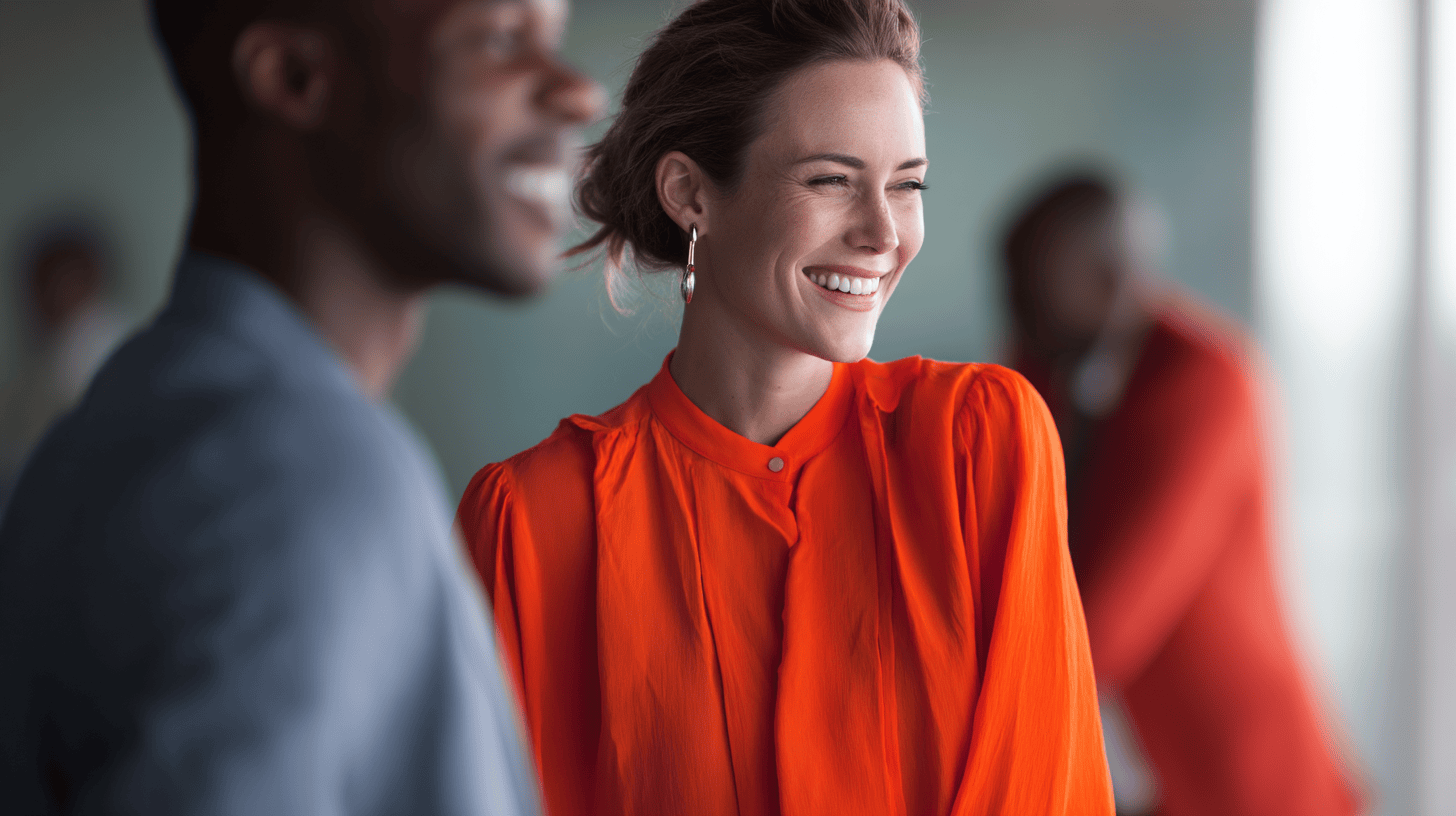 A woman in an orange blouse smiles brightly, engaging with a blurred group of people in a modern setting.