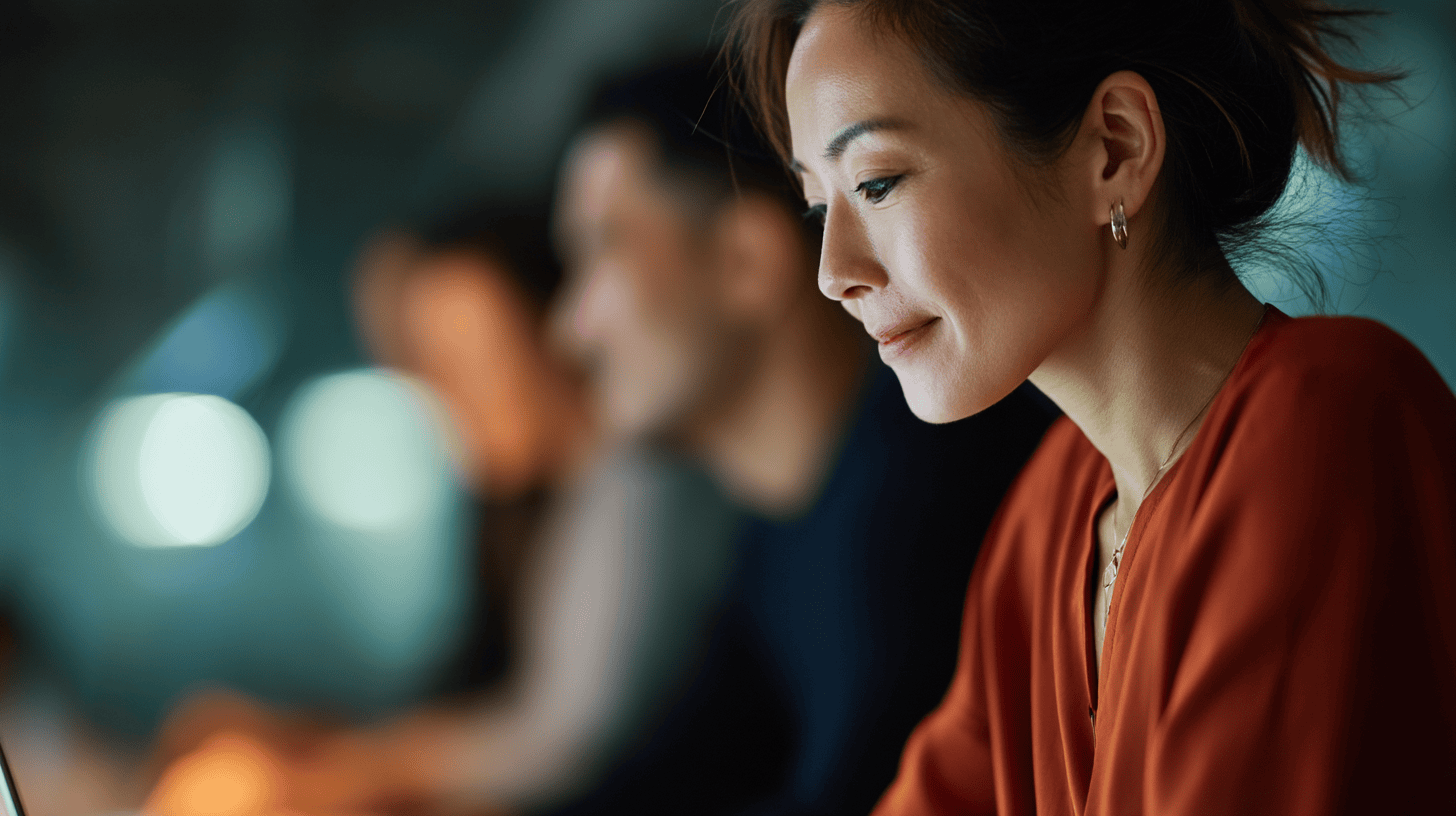 A woman in a rust-colored blouse focuses on a laptop, with blurred figures in the background. Soft lighting enhances the scene.