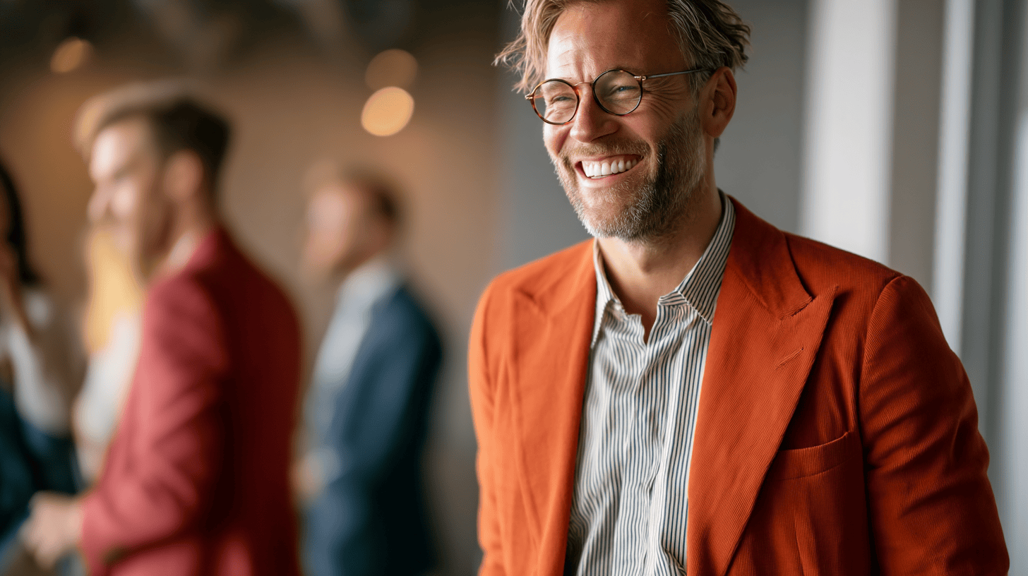 A smiling man in an orange blazer and striped shirt stands in focus, with blurred figures in the background.