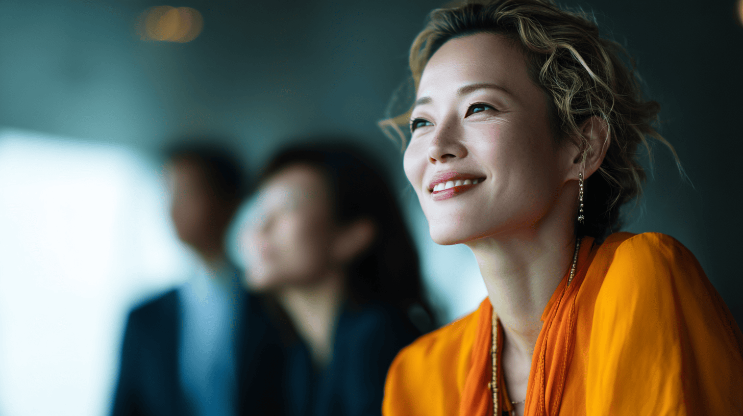A smiling woman in an orange top, with curly hair, is looking thoughtfully in a blurred background with two people.