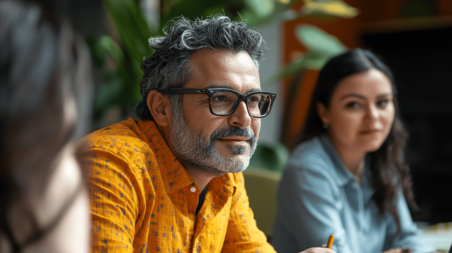 A man with curly hair and glasses smiles, wearing an orange shirt, while a woman sits nearby, observing attentively.