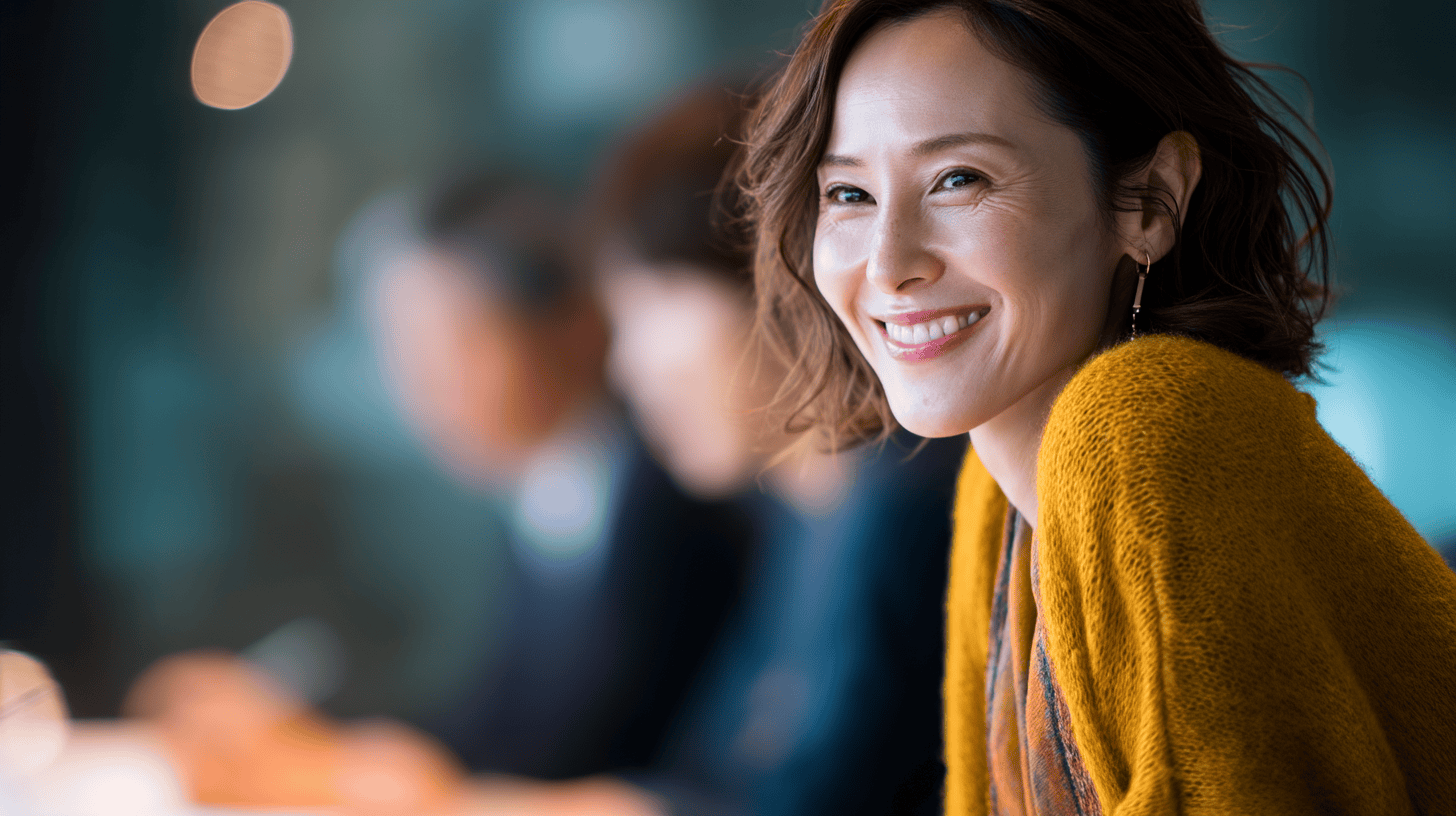 A smiling woman in a cozy mustard sweater, with blurred figures in the background, conveying a friendly atmosphere.