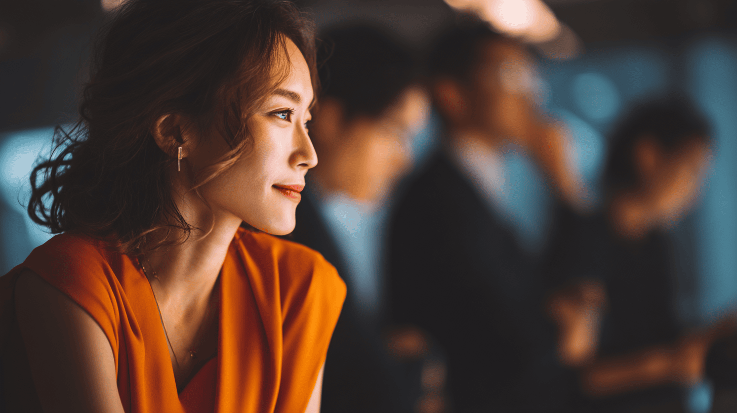 A woman in an orange top smiles softly, with blurred figures of people in business attire in the background.