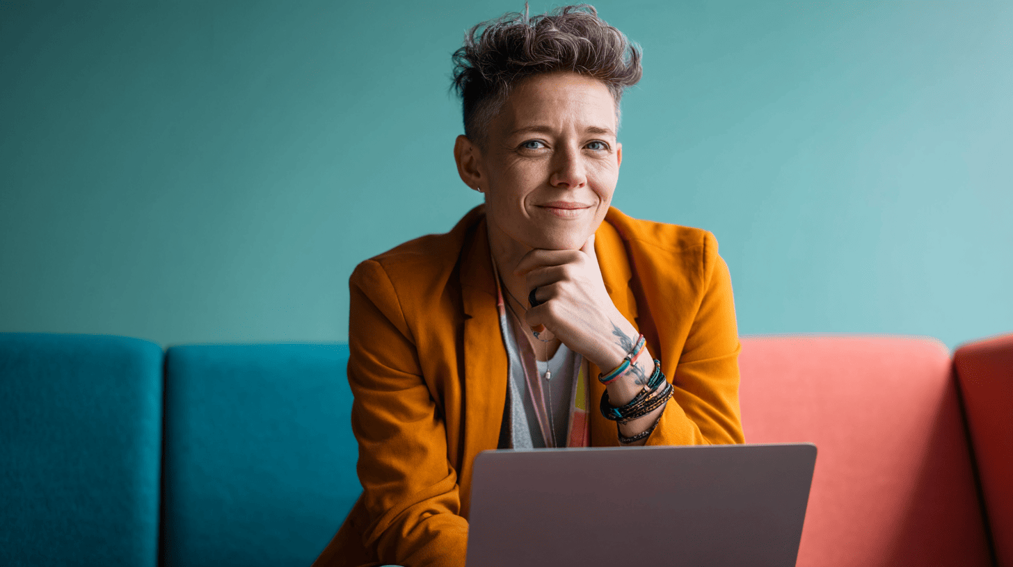 A person with short hair wearing an orange blazer sits on a colorful couch, smiling while looking at a laptop.