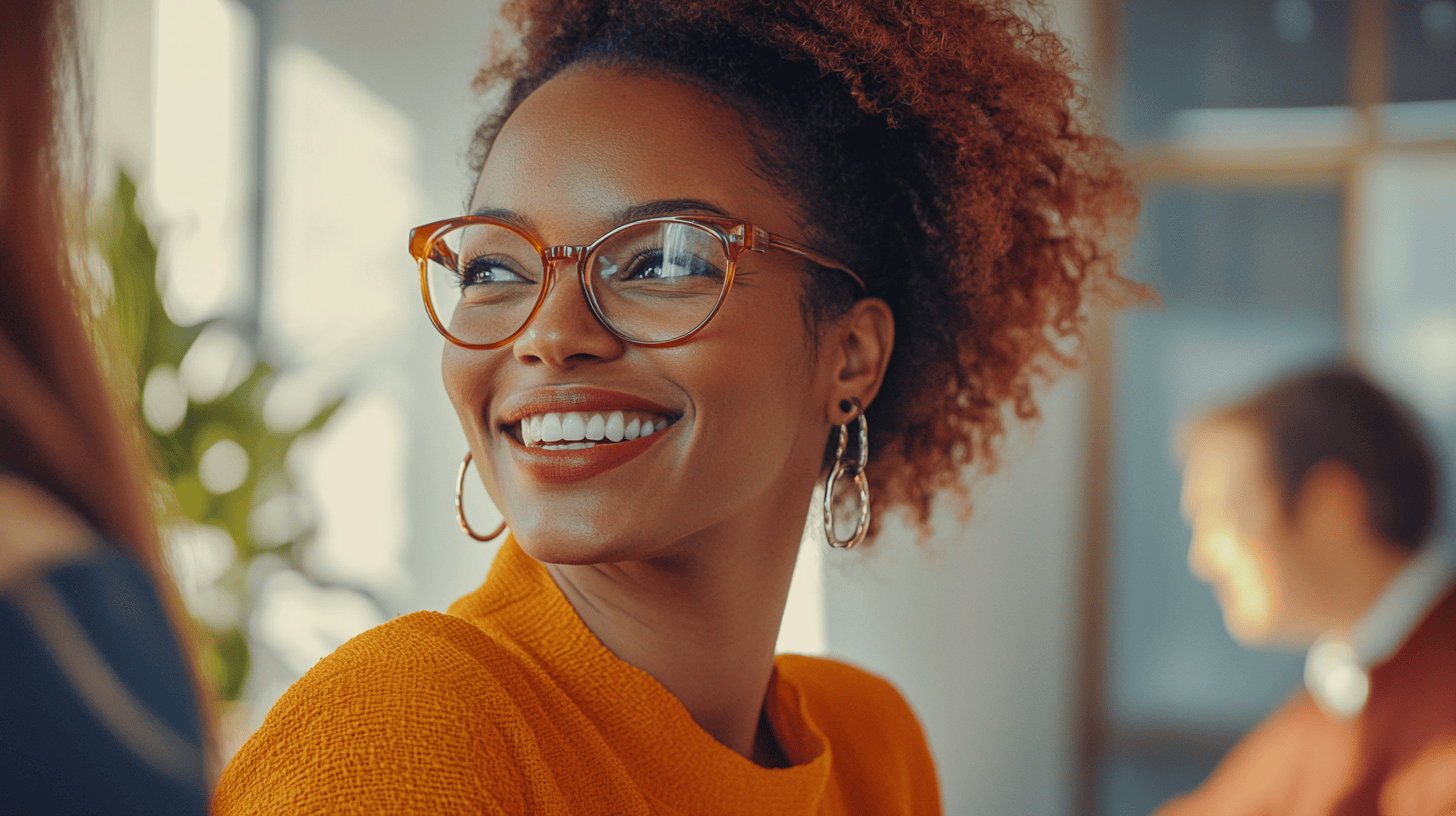 A woman with curly hair smiles brightly, wearing glasses and a textured orange sweater, in a softly lit indoor setting.