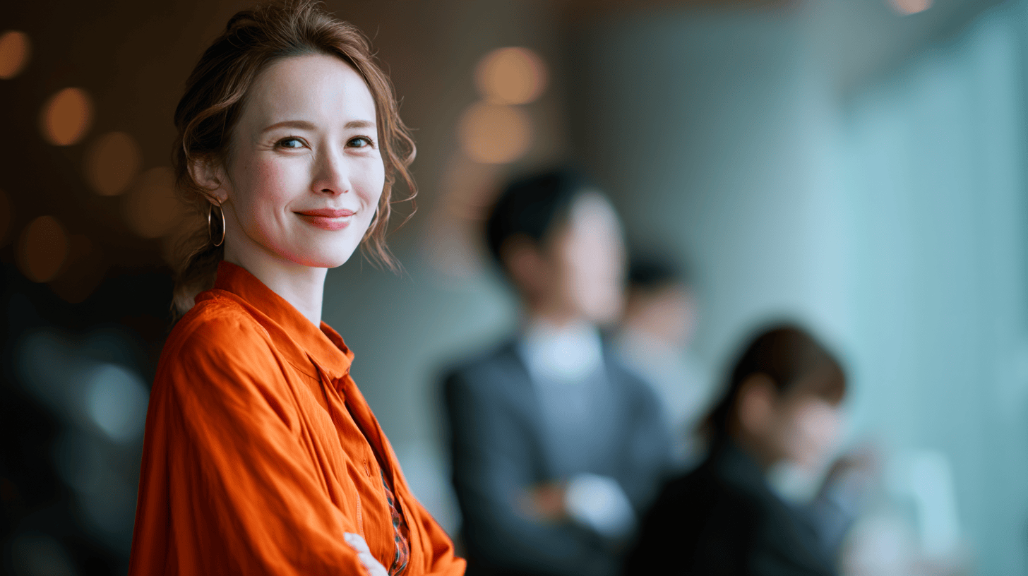A smiling woman in an orange shirt poses confidently, with blurred figures in the background. Bright indoor setting.