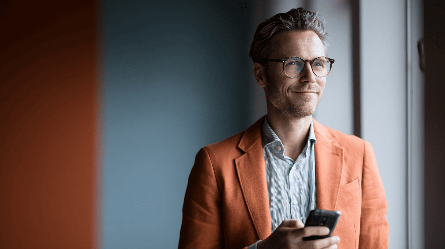 A man in an orange blazer stands by a window, holding a smartphone, with a soft smile and glasses on.