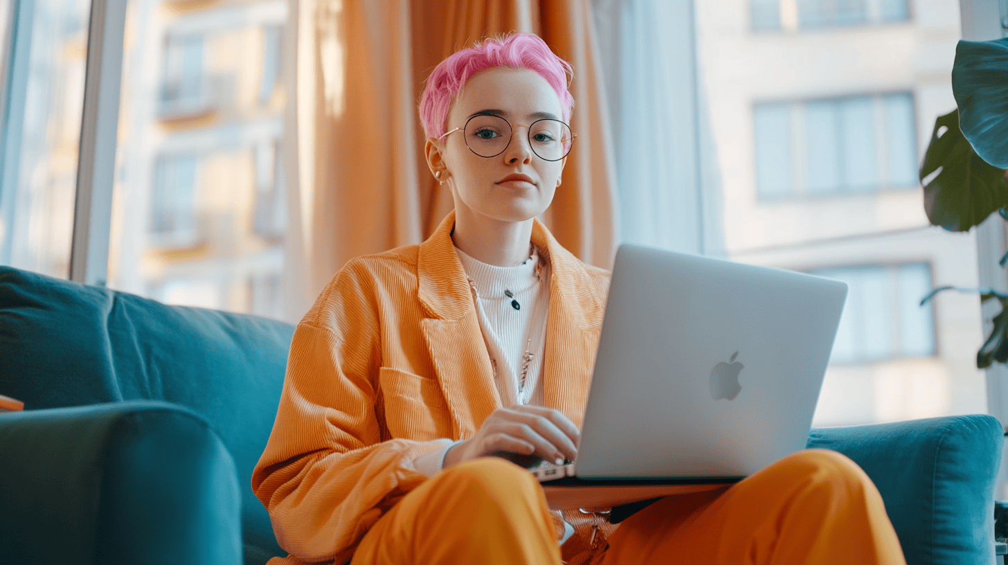 A person with pink hair and glasses sits on a teal couch, working on a laptop in a bright, modern room.