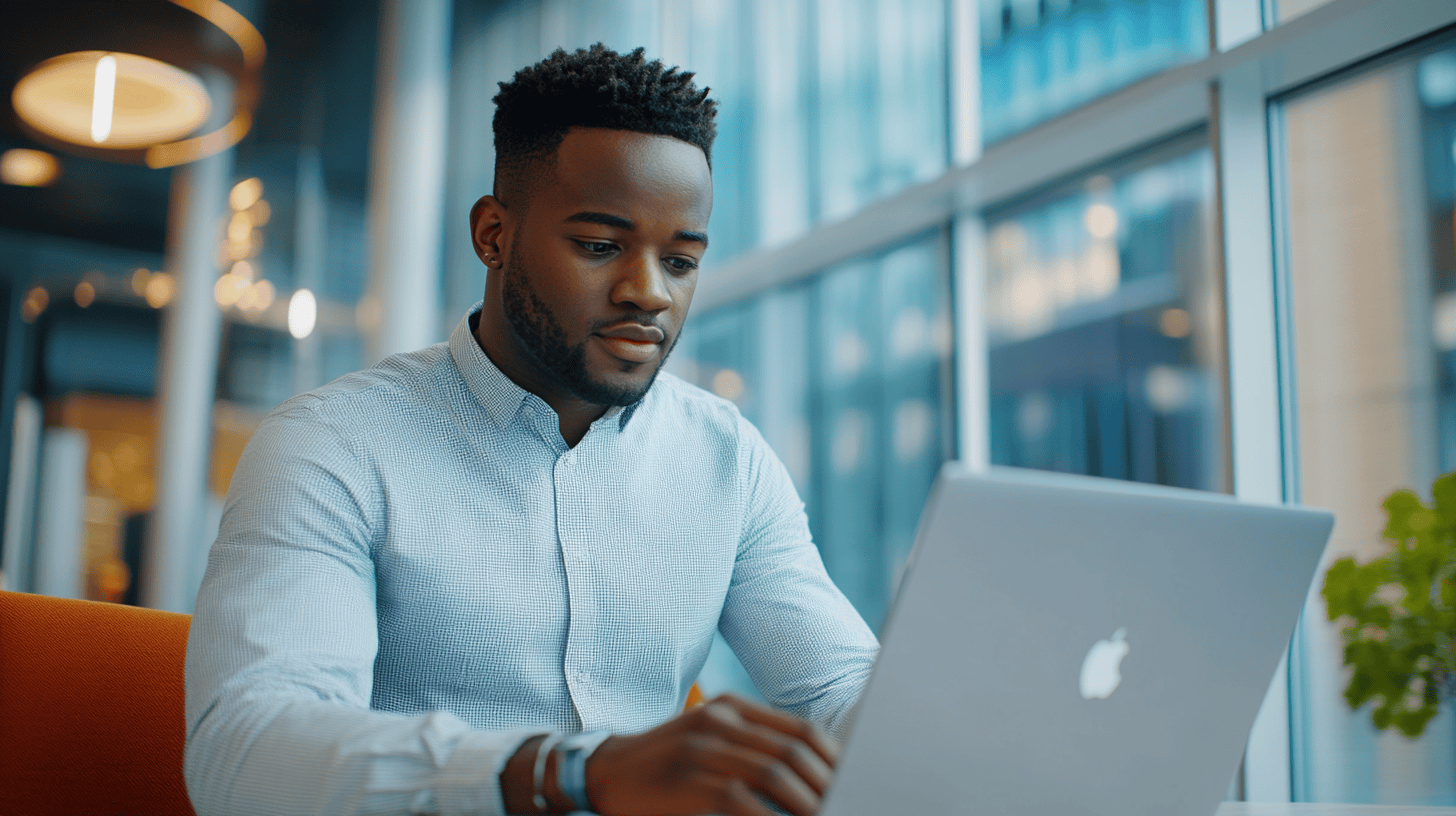 A young man types on a laptop in a modern office, with large windows and a plant in the foreground.