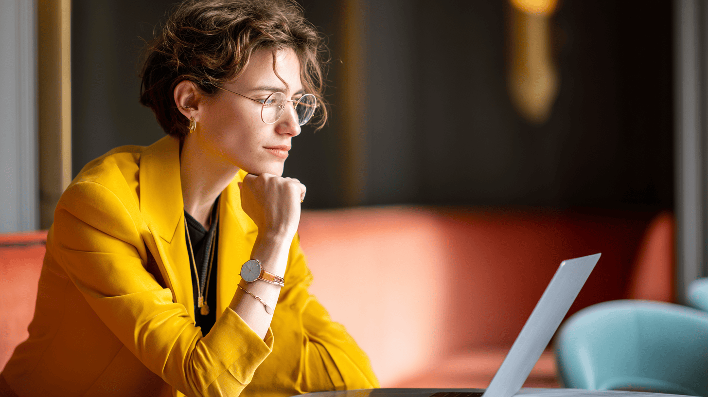 A person wearing a yellow blazer sits pensively near a laptop, with a colorful background of orange and blue.