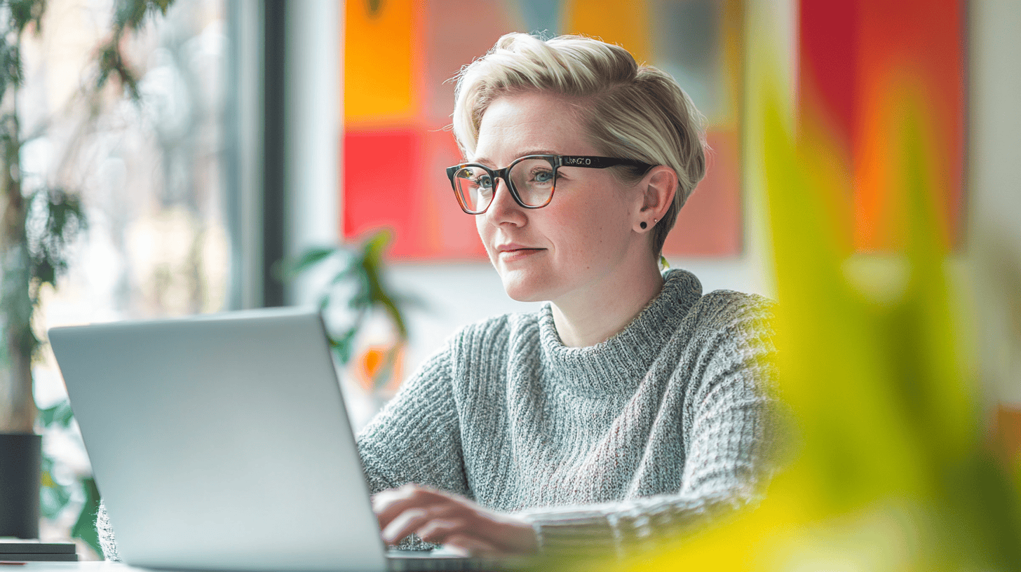 A woman with short blonde hair wearing glasses works on a laptop in a bright, modern space with colorful artwork.