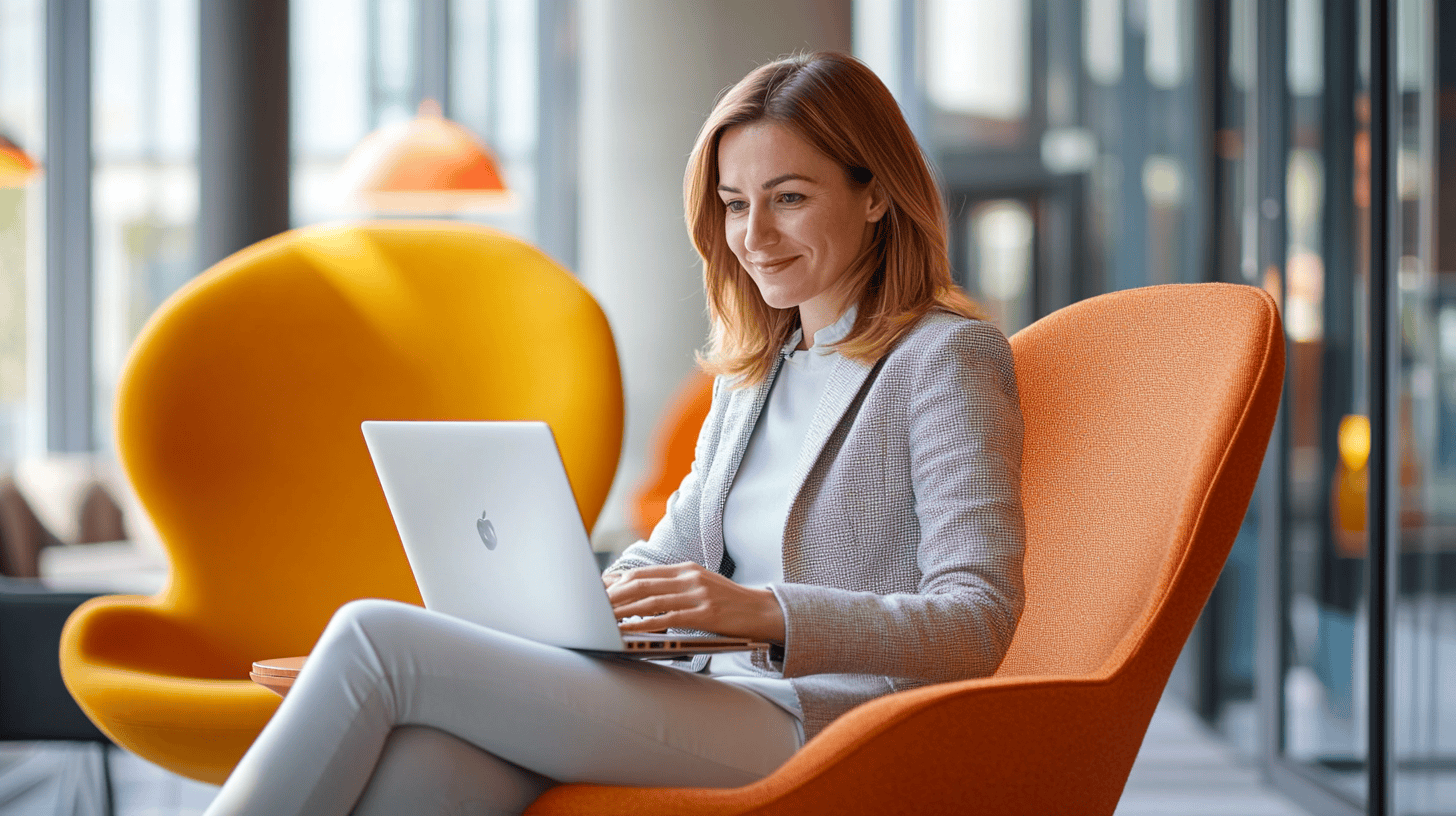 A woman sits in an orange chair using a laptop in a modern, bright office setting. Large windows provide natural light.