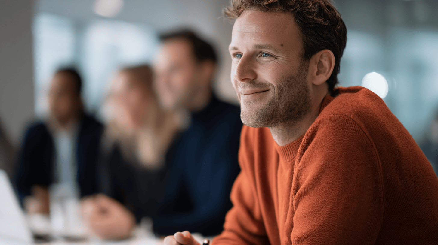 A smiling man in an orange sweater sits at a meeting table, with blurred colleagues in the background.