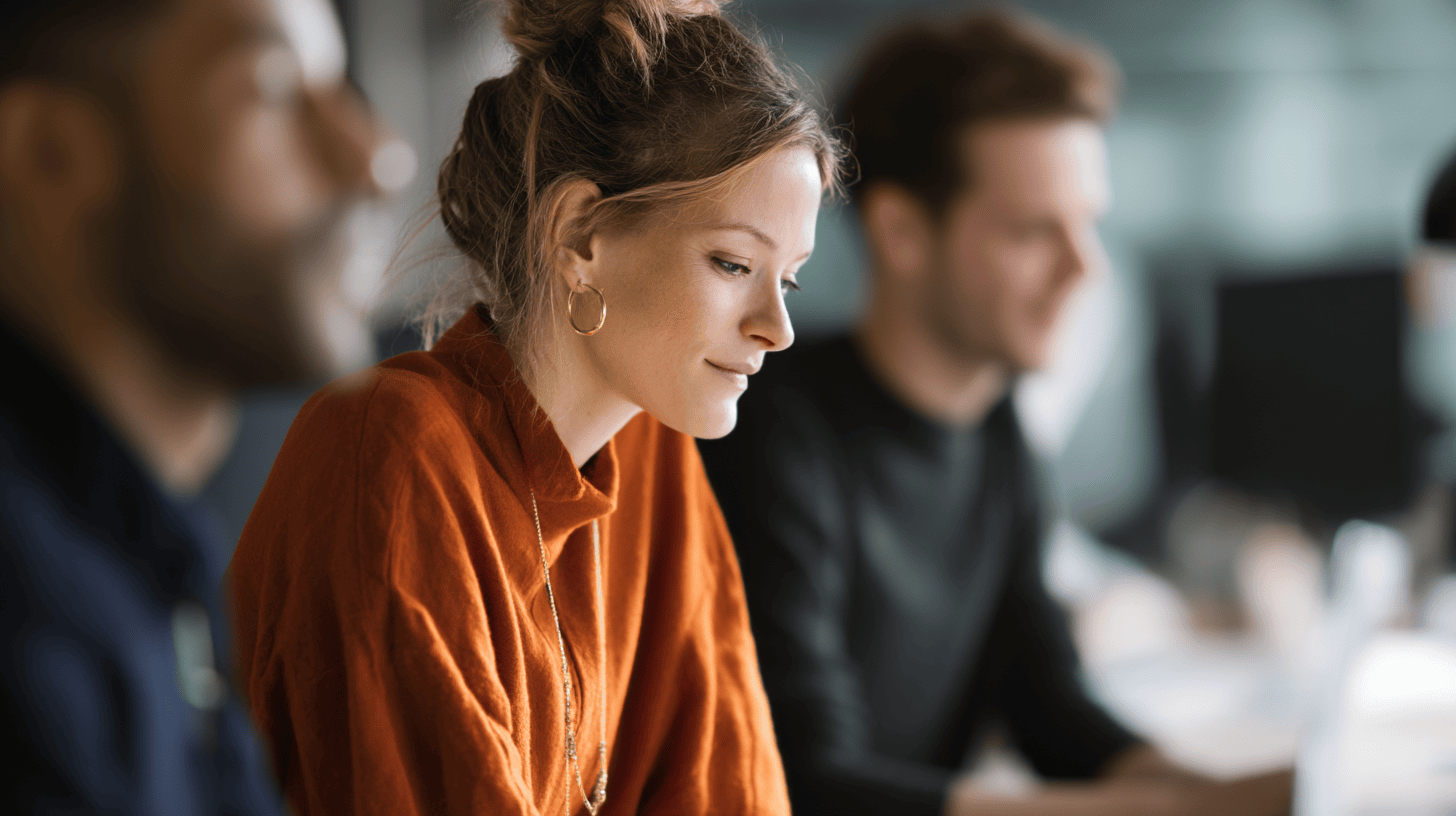 A woman in an orange sweater focuses on her work, with two colleagues partially visible in the background.