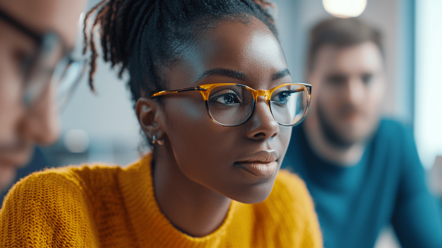 A close-up of a woman in glasses wearing a yellow sweater, focused on a task, with two blurred figures in the background.