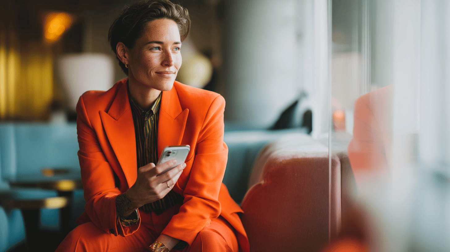 A woman in an orange suit sits by a window, holding a phone and looking thoughtfully outside.