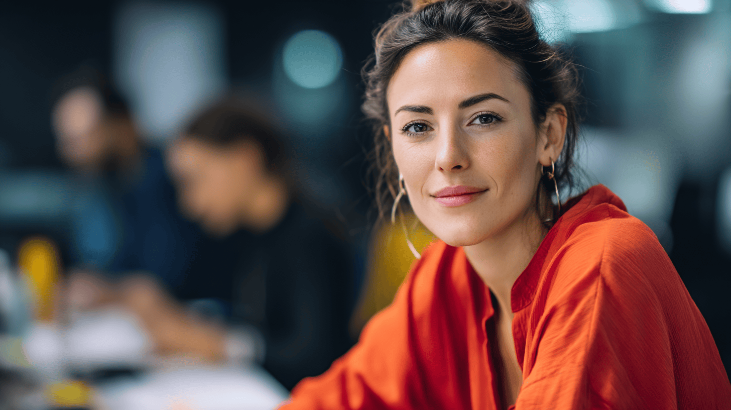A woman in an orange shirt smiles confidently at the camera, with blurred colleagues and work materials in the background.