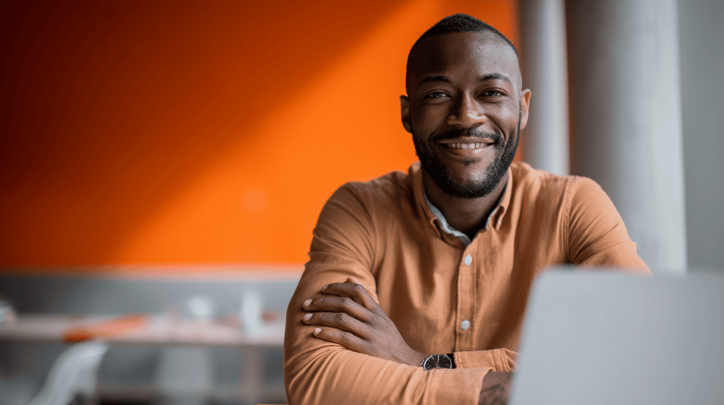 Smiling man with a beard in a brown shirt sits at a desk with a laptop against an orange wall.