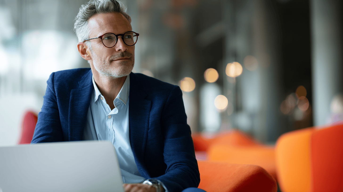 A middle-aged man with glasses sits at a laptop, appearing thoughtful in a modern, brightly colored seating area.
