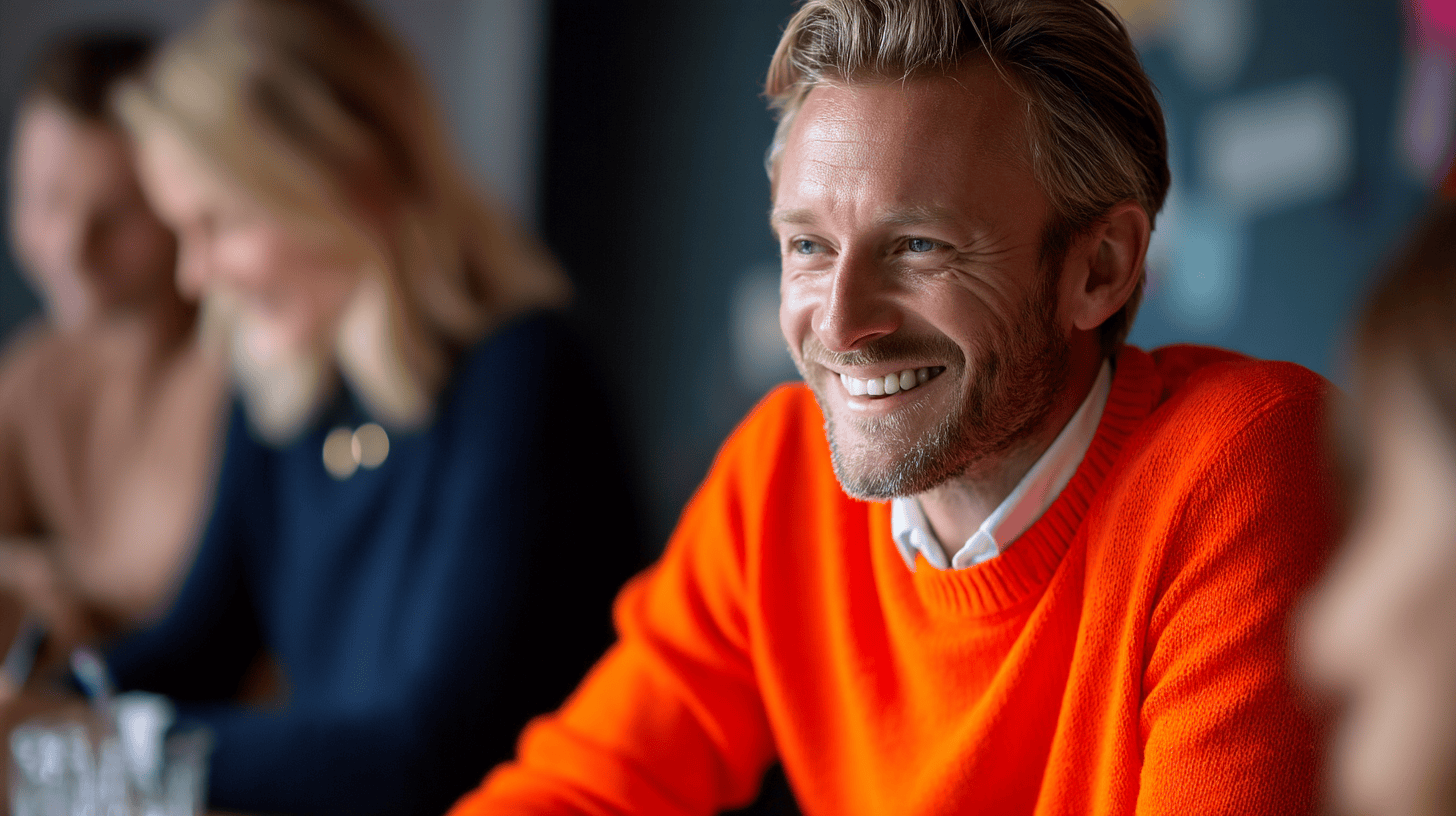 Smiling man in an orange sweater at a table with other blurred individuals, engaged in conversation. Soft focus background.